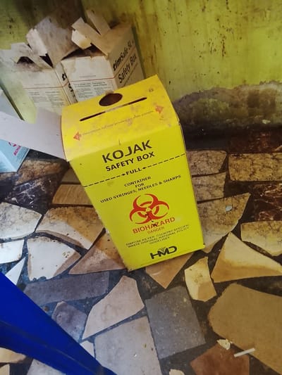 This image shows a close-up view of several biohazard disposal containers, primarily a yellow one labeled "KOJAK SAFETY BOX." The box is partially open and appears to be made of cardboard. It features prominent text indicating it is for "USED SYRINGES, NEEDLES & SHARPS," along with a biohazard symbol and the word "DANGER." Instructions for disposal are also visible, referencing "COUNTRY SPECIFIC WASTE DISPOSAL NATIONAL POLICY GUIDELINES."

To the left and behind the yellow box, other cardboard containers are visible, one of which is also a safety box, labeled "timSafe 5L Safety Box." A smaller blue box with the partial label "BIOHA" is also present.

The setting appears to be an indoor area with a rough, textured yellow wall in the background and a floor covered in broken tiles of various colors and textures, predominantly brown, beige, and black. The lighting suggests it might be daytime, but the overall environment appears somewhat unkempt. There are no people visible in the image, and no specific activity is taking place other than the arrangement of these discarded or stored medical waste containers. The image is focused on the objects and their labeling, emphasizing their purpose in medical waste management.