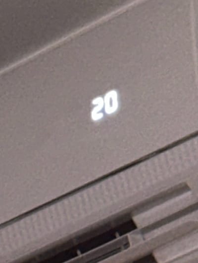 The image shows a close-up of a digital display showing the number "20" in white on a grey background. The display appears to be part of an electronic device, possibly an air conditioner or thermostat, given the textured grey surface and vent-like structures visible in the lower portion of the image. The lighting is somewhat diffused, creating a soft glow around the digits. There are no people, animals, or any other discernible objects or activities present in the scene. The background is a textured, neutral grey material. No specific location in Semenivka, Ukraine is identifiable from this image.