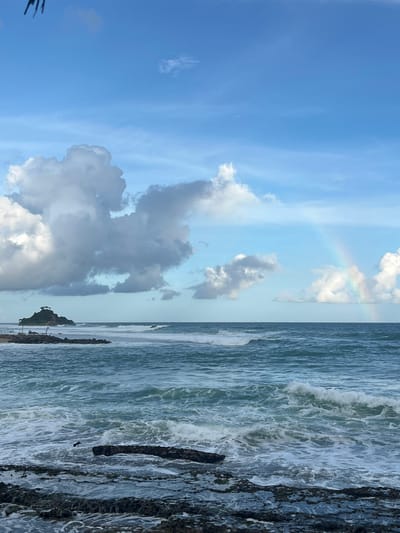 An overcast sky with dramatic clouds is visible above a turquoise ocean in Ahangama, Sri Lanka. A faint rainbow arcs in the distance. Waves crash against a rocky shore in the foreground. A small, tree-covered island with a structure on top is visible to the left, beyond a breakwater. A bird is in flight over the water.