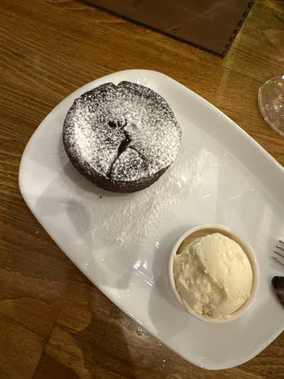 A close-up, overhead shot shows a dark chocolate lava cake dusted with powdered sugar and a scoop of vanilla ice cream on a white plate. The plate rests on a wooden table. The setting is likely a restaurant or cafe in Haskovo, Bulgaria, as indicated by the context provided.