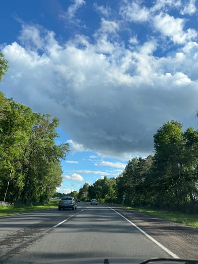 The image depicts a view from a vehicle driving on a paved road lined with trees on both sides. The road has a white line marking its edge. Several cars are visible on the road ahead. The sky is filled with a mix of dramatic, dark storm clouds and brighter, scattered white clouds against a blue sky. Sunlight filters through, casting shadows from the trees onto the road. The scene suggests a dynamic weather condition, possibly approaching or moving away from a storm. The lush green foliage of the trees indicates it's likely daytime during a warmer season. There is no visible text in the image. The setting appears to be a rural or semi-rural area in Belarus, given the location context.