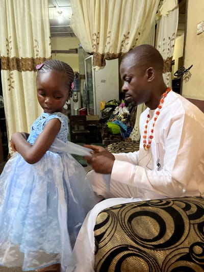 A man and a young girl are indoors. The man, facing left, is wearing a white shirt and an orange beaded necklace. He is adjusting the girl's light blue dress. The girl, also facing left, is wearing hair clips. The setting appears to be a home interior, with curtains and furniture visible in the background. There is no specific information in the image to verify the city of Rafin Bauna, Nigeria.