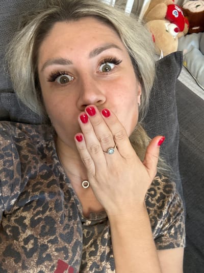 This is a selfie of a person with blonde hair, light skin, and green eyes, lying down and covering their mouth with their hand. They are wearing a leopard print shirt and a necklace with a circular pendant. Their fingernails are painted red, and they are wearing a ring on their finger. A stuffed animal is visible in the background. The image appears to be taken indoors, possibly in a home setting. The lighting suggests it could be daytime. The person's expression is somewhat obscured by their hand, but their wide eyes suggest surprise or perhaps playfulness. There is no visible text in the image. The location context provided is Novoaltaysk, Russia.
