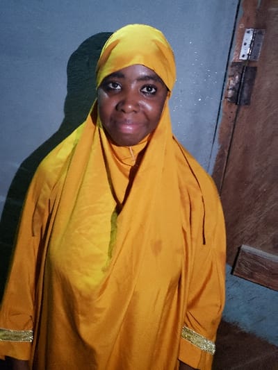 A young Black woman smiles wearing a bright yellow headscarf and gown with gold trim. The fabric drapes around her head and shoulders. She is indoors, with a plain wall behind her. Visible in the background are a brown door and some metallic hinges. The lighting in the room is soft and muted. The image is taken from a slightly high angle and rotated counterclockwise.