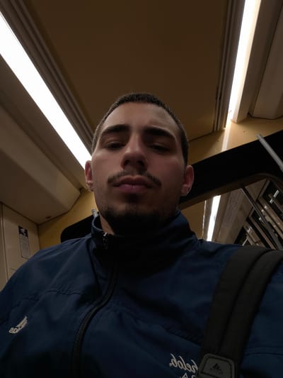 A young adult male is depicted in a close-up selfie, captured inside a public transport vehicle, likely in Hoenheim, France. The subject, positioned centrally, has short dark hair, a mustache, and a short beard, presenting a neutral facial expression. He wears a dark blue zip-up jacket with a white Adidas logo visible on the left chest. A black strap, consistent with a backpack, is visible over his right shoulder. The vehicle's interior features beige-colored overhead panels and walls, illuminated by elongated fluorescent light fixtures running along the ceiling. A white informational sign with black text and a pictogram is affixed to the wall behind the subject's left shoulder. Metal bars or framework are partially visible in the blurred background, suggesting window structures or internal partitioning of the vehicle.