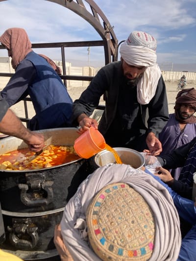 An outdoor scene in Chaman City Tehsil, Pakistan, depicts multiple individuals engaged in food and drink distribution from the bed of a vehicle, likely a truck, under a clear blue sky. In the foreground, a person is seen from behind, wearing a light grey head covering wrapped around an embroidered cap with colorful square patterns. Their hands hold a metal bowl.
To their left, a large, dark-colored metal pot containing a red-orange stew with visible chunks of potatoes and meat is being stirred by a man partially obscured, wearing a blue vest and a light brown head covering. Central to the image, a bearded man wearing a white turban over a silver-embroidered cap and a black tunic is pouring an orange liquid from an orange plastic measuring cup into a metal bowl held by the foreground individual. His hands bear rings.
To the right of the central man, another individual wearing a dark brown turban and a purple garment observes the pouring action. Multiple hands are visible, suggesting more recipients or helpers. The large cooking pot rests on another identical pot, creating a tiered cooking station within the vehicle's metal frame.
The background reveals a dusty, arid landscape with concrete block walls and distant mountains under bright sunlight. A person on a motorcycle is visible further in the background.