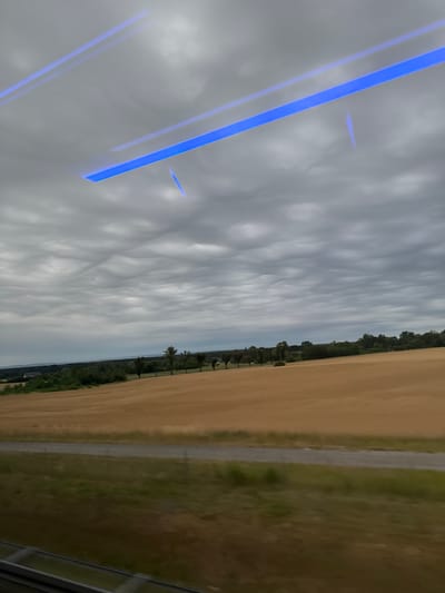 The image depicts a landscape scene, likely taken from a moving vehicle, possibly a train, given the blurred foreground and the reflections of blue light strips on the window. The sky is overcast with distinct, rippling cloud formations. In the middle ground, a field of dry, golden crops stretches horizontally, bordered by a line of trees and foliage. A narrow dirt road or path runs through a strip of green grass in the foreground. The background shows a distant horizon with hints of more vegetation and possibly buildings. The location is identified as Brzezie, near Sulechowa, Poland. There are no people or animals visible in the frame. The overall impression is one of rural scenery under a dramatic sky.