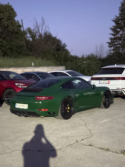 In Arbanassi, Bulgaria, a dark green Porsche 911 is parked on an asphalt surface. The car has a license plate that reads "PB 2090 CE." To the left of the Porsche, a red car is partially visible. Behind the Porsche, a white Audi SUV is parked, displaying a license plate that reads "856 M 306." The scene is set against a backdrop of trees and a clear sky, suggesting it is either early morning or late evening. A shadow of a person holding a phone is cast on the ground in front of the Porsche.