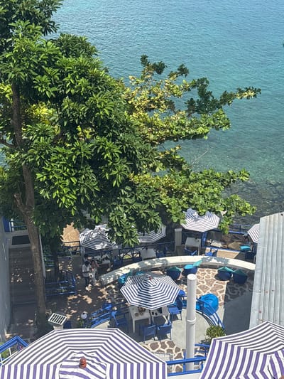 Viewed from above, the image shows a section of Kamala, Thailand. Part of the turquoise ocean is visible on the left, separated from a waterfront patio by lush green trees. The patio area, constructed with white concrete and blue railings, features blue and white striped umbrellas shading tables and chairs. Some people are seated at the tables under the umbrellas. The overall atmosphere appears relaxed and vacation-oriented.