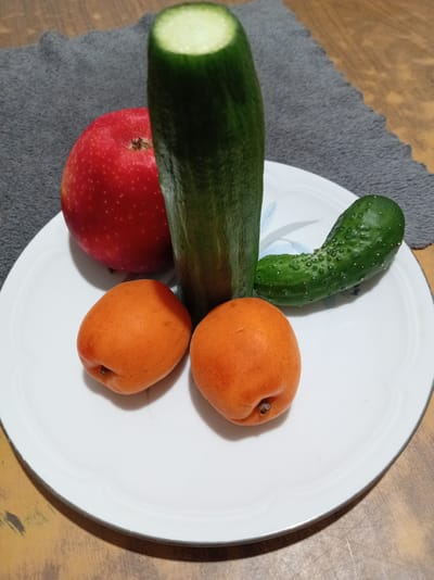 A white plate rests on a wooden surface, partially covered by a gray cloth. The plate contains several pieces of fruit and vegetables: a bright red apple, a whole cucumber, a small pickle, and two apricots. The arrangement of the items on the plate is suggestive. The scene is indoors. There is no information about the city of Bugulma, Russia.