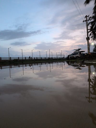 The image shows a flooded street or walkway with reflections of a cloudy sky, lampposts, a railing, palm trees, and a car. A utility pole with wires is visible on the right side of the frame. The sky is a gradient of pale blue and pink hues, suggesting either dawn or dusk. A few silhouetted figures can be seen on the walkway behind the railing. The water appears murky, and debris is visible near the utility pole. The scene is likely in a coastal area due to the presence of palm trees. The specific location in Badem, India cannot be determined from the image alone.