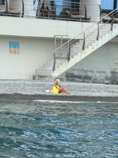 A person in a yellow top and white shorts is sitting on a pebble beach next to a body of water. The person has blonde hair and is wearing white shoes. The water appears to be a light blue color with ripples on the surface. To the left of the beach is a large, light-colored building with a set of stairs leading down to the beach. The stairs are made of stone and have metal railings. The building has several windows and a modern design. A small, colorful artwork is visible on the wall of the building. The scene appears to be outdoors during the daytime, with natural light illuminating the area. The overall impression is one of a relaxed and leisurely setting, possibly a beach resort or a public recreational area. The orientation of the image is sideways.
