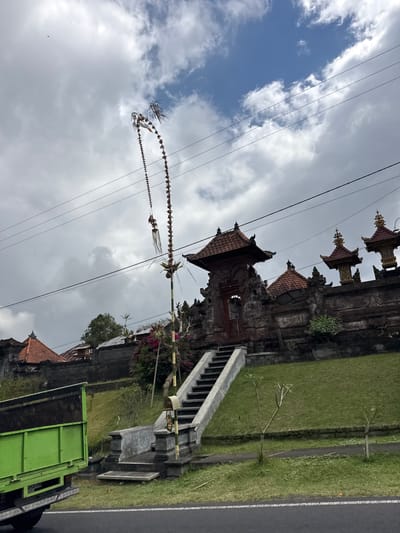 This is a vertical, eye-level shot of a street scene in Kintamani, Indonesia. The sky is overcast with dramatic, heavy clouds. In the foreground, a large, green truck is parked on the right side of the frame, facing away from the camera. The truck's bed is empty. Adjacent to the truck, a stone wall and pathway lead up to a traditional Balinese temple complex. The temple features intricate stone carvings and multi-tiered roofs topped with gold.
A long, ornate bamboo pole, a Penjor, stretches diagonally across the sky, adorned with festive decorations, including what appear to be flowers and woven palm leaves. This is a common sight in Bali during holidays and celebrations. The Penjor is suspended by utility wires that also cross the frame.
To the left of the temple, a well-maintained green lawn slopes gently upwards. The scene appears to be a public area, possibly near a road, given the asphalt surface visible on the far right with a white line marking the edge. The overall impression is one of a cultural and everyday scene in Bali, set against a backdrop of a cloudy sky.