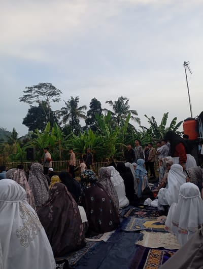 The image shows a group of people gathered outdoors in a clearing surrounded by trees and foliage. They are all wearing traditional Islamic clothing and some are kneeling, praying, or sitting on the ground. There are a few prayer rugs spread out, and the people are facing towards the left of the frame. The scene is set in an open-air area with a lush green background, possibly a mosque courtyard or a garden. There is a blue plastic tarp covering the ground. The overcast sky suggests it is early morning or late afternoon. The location based on the provided coordinates is in the village of Kutawaringin, Indonesia.