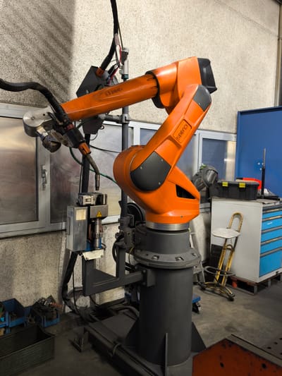 An industrial robotic arm, painted bright orange, is the primary subject in this indoor image. The robot is mounted on a dark grey cylindrical base and appears to be a welding robot, as evidenced by the welding torch assembly attached to its end effector. The robot's arm segments are articulated and show the "CLOOS" brand name.

The background consists of a textured, light-colored wall with a prominent shadow cast across it, suggesting light sources from the side and above. To the left of the robot, there is a large window with a metal door visible behind it. The window frames are grey.

In the mid-ground and right side of the image, there is a blue metal cabinet with drawers, a black toolbox with yellow latches, and a yellow metal stool with a round seat. A metal ladder leans against the cabinet. Several blue plastic bins and a metal container holding various tools and components are visible on the floor in the lower left. The floor appears to be concrete.

The scene is likely a workshop or factory setting, possibly related to manufacturing or metal fabrication. Given the context provided about the city of Bad Honnef, Germany, this scene could be located in a production facility or a vocational training center within that city. There are no people visible in the image. The lighting is consistent with artificial workshop lighting.