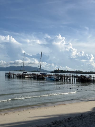 The image depicts a scene at the Baan Bang Rak pier in Thailand. The foreground shows a sandy beach with gentle waves lapping the shore, casting shadows from unseen objects onto the sand. In the mid-ground, a wooden pier extends into the calm, blue water. Several boats are moored at the pier, including two sailboats with tall masts and a white motorboat. Beyond the pier, a chain of buoys floats on the water's surface. In the background, lush green hills rise against a sky filled with dramatic, white cumulus clouds. The overall atmosphere is serene and picturesque, characteristic of a tropical coastal setting.