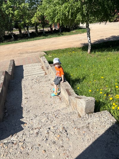 A young child lies on a concrete surface in Ферма, Russia. The child wears an orange shirt, gray shorts, a light-colored hat, and blue sandals. The concrete surface has a gravelly texture and is casting a large shadow. To the left is a dirt road bordered by trees and a fence. To the right is grass with yellow flowers. The image appears to be taken from an elevated position.