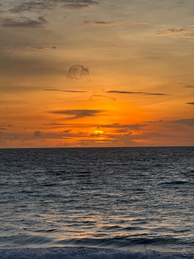 The image depicts a sunset over the ocean, presumably off the coast of Pecatu, Indonesia. The upper half of the frame is filled with a sky transitioning from a soft grey-blue at the top to vibrant orange and yellow hues near the horizon, where the sun is visible as a bright, glowing disc partially obscured by a horizontal band of clouds. Scattered, elongated dark grey clouds are present across the sky, with a distinct, somewhat circular cloud formation visible in the upper left quadrant. The lower half of the frame shows the dark blue-grey surface of the ocean, exhibiting subtle wave patterns and ripples that reflect the sky's colors. The horizon line is sharply defined and horizontal. At the very bottom of the image, the white foam and crests of breaking waves are visible in the immediate foreground. There are no people, man-made objects, or specific actions/interactions depicted in the scene.