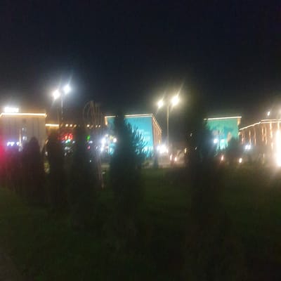 The image depicts a nighttime scene in Turkistan, Kazakhstan. Two large, rectangular, aquamarine-colored billboards are illuminated by spotlights. Streetlights illuminate the area and there appear to be buildings decorated with lights, indicating an urban environment. Some foliage is present in the scene.