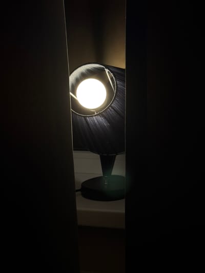 A lamp with a black shade is illuminated in a dark room, as viewed through a narrow horizontal opening. The light source is a bright white bulb. The lamp appears to be mounted on a wall, with the mounting hardware partially visible. The environment is predominantly dark, with the lamp being the primary source of illumination. It is difficult to determine the location as being in Rossiyskiy, Russia based on the provided image.