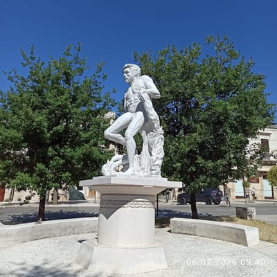 The image shows a white marble statue of a muscular man in a dynamic pose, possibly wrestling with a creature or draped in fabric. The statue is mounted on a multi-tiered, octagonal pedestal, which sits on a circular base paved with small, light-colored stones. Two large, leafy green trees flank the statue. In the background, there is a street with a parked dark-colored SUV and several people walking. A building with classical architectural elements, including arched windows and a balcony, is visible behind the trees. The sky is clear and blue. The scene is likely a public square or park in the city of Ruvo di Puglia, Italy, given the architectural style and vegetation. A timestamp "06.07.2026 09:40" is superimposed on the lower right portion of the image.