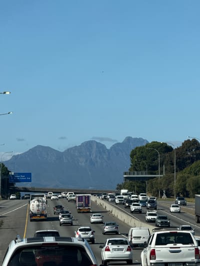 This image captures a busy highway scene, likely near Bellville, South Africa, as indicated by the visible road names and vehicle registrations. The photograph appears to be taken from an elevated perspective, looking down a multi-lane road experiencing heavy traffic.
Numerous vehicles, including cars, vans, and trucks, are visible across all lanes, suggesting either a traffic jam or slow-moving congestion. In the foreground on the right, a prominent white Toyota Hilux bakkie is seen, bearing the text "TOYOTA," "HILUX," and the license plate "CJ 56068." A smaller sticker on its rear reads "STEVES AUTO CLINIC MILNERTON." Further down the right lane, another car has the license plate "CF 218-558." On the left lanes, a tanker truck with "000" on its side is part of a queue of vehicles.
Overhead streetlights line the highway, and a large blue road sign is visible on the left. The sign provides information for upcoming exits, stating "23 R302," "Durban Road," "W van Schoor Ave," and "1km."
The background reveals a majestic range of rugged mountains under a clear, bright blue sky, typical of the Western Cape landscape. Lush green trees grow along the side of the highway, bordering the distant mountains. The scene is illuminated by strong daylight, indicating sunny and clear weather conditions. The overall impression is one of a bustling urban or suburban transport corridor during peak hours.