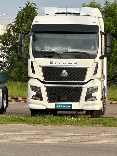A white Sitrak heavy-duty truck is presented in a direct frontal view, parked outdoors in Бараўлянскі сельскі Савет, Belarus. The truck features a prominent black grille with a hexagonal Sitrak emblem centrally located in the upper section, and the word "SITRAK" in silver capital letters positioned above it. A lower teal-colored plate on the front bumper also displays "SITRAK" in white lettering. Integrated headlights with distinct LED daytime running lights are visible on both sides of the lower fascia. Large black side mirrors extend from the A-pillars. A black sun visor is situated above the windshield. The cab's roof exhibits white-colored structural elements and what appear to be multiple small antennas.
To the immediate left of the truck, a portion of a silver wheel with a chrome hubcap from another vehicle is visible. The ground directly in front of the truck's bumper is a narrow strip of green grass dotted with small white flowers. Below this, a grey paved surface with a textured, rectangular pattern, consistent with pedestrian paving or a sidewalk, is present, leading to a dark asphalt road surface in the foreground. In the background, behind the truck, numerous green deciduous trees are densely arrayed, partially obscuring distant light-colored buildings on the left. Wooden pallets and yellow and black striped ground barriers are visible on the ground behind the truck. The scene is illuminated by natural daylight. No individuals are present in the image.