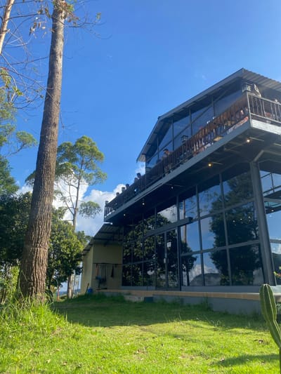 The image shows a modern, glass-fronted building with a balcony on the upper level. The building is located on a grassy slope with a large tree to the left. The balcony is crowded with people, some of whom are standing and some are sitting. The building is surrounded by lush vegetation. The sky is clear and blue. The building is located in the city of Kecamatan Tawangmangu, Indonesia, which is situated at the coordinates: Latitude -7.661, Longitude 111.185.  The large glass windows are reflecting a view of the surrounding landscape.  There is a sign on the entrance of the building.  A cactus is in front of the building.
