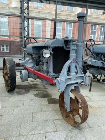 The image shows a vintage gray tractor on display at the Museum Complex of Military Equipment "Ural Military History" in Verkhnyaya Pyshma, Russia. The tractor, likely a historical exhibit, is positioned on a stone-tiled outdoor area with a glass-enclosed structure in the background, possibly part of the museum. The tractor has large, spoked metal wheels, a prominent engine area with a radiator and headlight, and a steering wheel visible. A red bar is affixed to the front chassis. The overall condition of the tractor suggests it is a restored or preserved piece of machinery. The background reveals a building with multiple windows and architectural detailing.