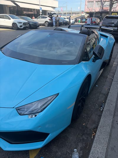 The image shows a light blue sports car parked on a city street. The car is positioned at an angle, with its front right corner prominently displayed. The street appears wet, suggesting recent rain or cleaning. Cigarette butts and a discarded can are visible on the ground near the car, indicating a somewhat unkempt urban environment.

In the background, other vehicles are parked in a line, including a black SUV and several white cars. Some people are standing near these vehicles, appearing to be engaged in conversation. The architecture in the background suggests an urban setting with storefronts and buildings. A traffic light and street signs are also visible, further reinforcing the urban context. One sign displays a "P" for parking.

The time of day appears to be daytime, with clear, bright lighting casting shadows. The weather seems to be fair, despite the wet street. The overall impression is of a casual urban scene where the sports car is a striking focal point amidst the everyday activity of the city. No specific event is clearly depicted, but the presence of people and parked cars suggests a normal day in Zurich, Switzerland.

Visible text includes "TAXI" on a black vehicle in the background.