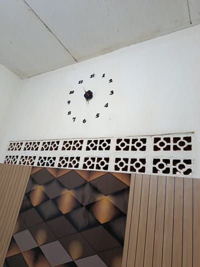 The image shows an interior wall in Depok, Indonesia. A white ceiling is visible at the top of the frame, with a slight discoloration and a visible seam. The wall below is painted white and features a large, modern wall clock with black numbers and hands. The clock's hour hand is pointing between 10 and 11, and the minute hand is pointing between 4 and 5. Below the clock is a row of decorative white concrete ventilation blocks with a circular pattern. Beneath the ventilation blocks, the wall is adorned with a visually striking wallpaper or paneling. This lower section consists of two distinct patterns: on the left, a geometric design of diamonds in shades of brown, gold, and black, with a reflective, almost metallic finish; and on the right, vertical wooden planks in a light brown color with thin, dark vertical lines. The scene appears to be a domestic or commercial interior space.