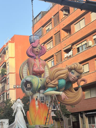 In Alicante, Spain, a large, colorful sculpture is being erected in an urban setting. The sculpture features two exaggerated, cartoonish female figures. The upper figure has pink hair and wears a red and gold patterned top, with a blue and white patterned ceramic pot balanced on her head. She is suspended by a crane. Below her, a blonde figure with blue eyes is positioned in a reclining pose, wearing a green dress. A ladder leans against the sculpture, suggesting ongoing assembly or maintenance. In the background, multi-story brick buildings with balconies and air conditioning units are visible. A white truck with lettering is partially seen on the left side of the frame, and a sign for "SUPERMERCADO ES" is visible on a building in the lower right. The scene appears to be taking place during daylight hours, with a clear blue sky.