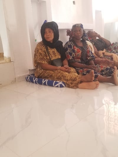 Three adult females are seated barefoot on a white tiled floor in an indoor setting within Edu Town, Nigeria. A fourth person's head and shoulder are partially visible on the far right. The female on the left wears a black headscarf, a brown and yellow patterned dress, and carries a black shoulder bag; she is seated on a blue and white patterned mat or cushion, looking slightly to her right with hands clasped. The female in the center wears a blue, white, and red patterned headwrap and a matching dress with dark embellishments, looking directly forward with hands clasped. The partially visible female on the right wears a patterned garment and appears to be resting with her head tilted back. The room features plain white walls with built-in niches or shelving.