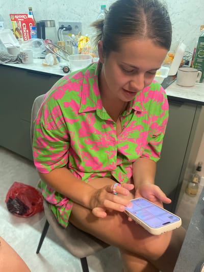 A young woman with brown hair tied back sits on a chair, looking down at her phone. She wears a green and pink floral print shirt and has a ring on her finger. The background shows a kitchen counter cluttered with various items, including food packaging, bottles, and containers. A red plastic bag is visible on the floor to her left. The overall scene suggests a casual indoor setting, possibly a home kitchen. The location of Novorossiysk, Russia is not discernible from the image itself.