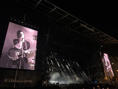 The image captures a concert at night in Porto, Portugal, likely during the Primavera Sound festival, as evidenced by the "Estrella Damm" branding visible on banners and the stage structure. The scene is dominated by a large stage featuring prominent video screens displaying a black and white image of a musician playing a guitar. The stage is equipped with elaborate lighting rigs and speakers. Smoke or fog machines are active, creating atmospheric plumes illuminated by spotlights. In the background, a crowd of concert-goers can be seen, their hands raised. The overall atmosphere is that of a large-scale outdoor music event.