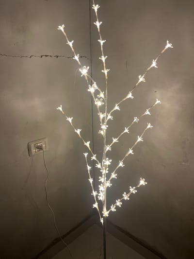 A lit faux floral branch is mounted against a neutral-toned wall. The branch has numerous small, white flower-shaped lights along its length. The wall is visibly textured, with a vertical crack extending upwards. An electrical outlet is visible near the base of the wall, with the branch's power cord plugged into it. The overall scene is illuminated by the warm light emitted from the decorative branch.