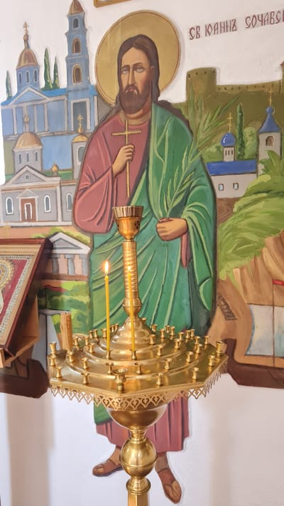 This media file captures an interior scene, likely within a church or religious building, given the presence of a fresco and a large, ornate brass candelabrum. The fresco depicts a religious figure, possibly Saint John the Baptist, with a halo, holding a cross and a palm frond. Behind him, stylized depictions of buildings, perhaps churches or monasteries, are rendered in blues, grays, and browns. The figure is dressed in a green robe and a reddish-brown tunic.

The candelabrum is the dominant object in the foreground. It is made of polished brass and features multiple arms, each designed to hold a candle. Two candles are visibly lit, casting a warm glow. The intricate metalwork of the candelabrum suggests it is a significant decorative and functional item within the space.

The overall impression is one of solemnity and devotion. The lighting appears to be a mix of ambient light and the warm glow of the candles, suggesting it might be evening or a dimly lit part of the day. The fresco style is characteristic of Eastern Orthodox art. Visible text in the upper left corner appears to be in Cyrillic script, reading "СВ. ІОАННА СОЧАВСЬКОГО," which translates to "Saint John of Sochi." This confirms the identity of the figure in the fresco and provides a specific cultural context. The location is identified as Bilhorod-Dnistrovskyi, Ukraine.