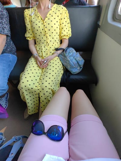 The image shows a top-down view of two people sitting on a dark-colored bench, likely within a public transportation vehicle, possibly a train or bus, as indicated by the interior seating and window framing on the right. The person on the left is wearing a dark, patterned shirt and blue jeans, with one foot visible, wearing a grey and blue athletic shoe. To the right of this person sits another individual wearing a yellow dress with a pattern of dark hearts. This person has their hands clasped in their lap, and is wearing a watch on their left wrist. A patterned blue bag is resting next to them. In the foreground, the legs of a third person are visible, wearing pink shorts and a pair of reflective, blue-tinted sunglasses are placed on top of the shorts. A light blue strap, possibly from a bag, is visible to the left in the foreground. The environment appears to be dimly lit, with the interior of the vehicle being the primary focus. The location is described as Shchomyslitski Sel'ski Savet, Belarus, which suggests an area near Minsk.
