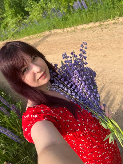 A young woman with dark auburn hair and bangs, wearing a red dress with a white floral print, is holding a bouquet of purple lupine flowers. She is standing on a dirt road, with green foliage and more lupine flowers in the background. The location is in the village of Garevaya, Russia. The image is a selfie, taken outdoors during daylight hours.