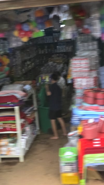A cluttered store in Pơng Drang, Vietnam is filled with stacked plastic containers of various colors (red, blue, green, yellow) and assorted merchandise. A person wearing a dark shirt and shorts stands amidst the merchandise, near a green object. Shelving units display fabrics and other goods.
