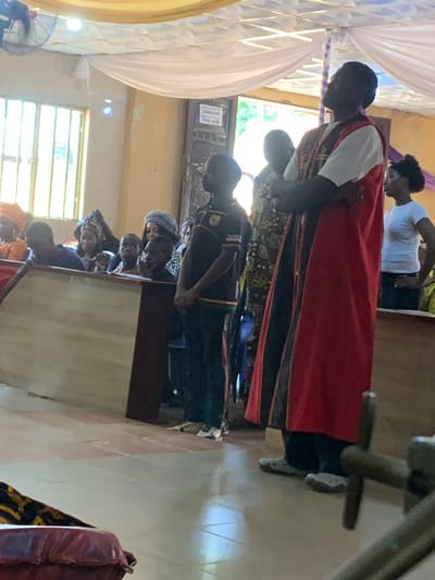 A group of people is seen indoors. A man in a white shirt and red robe stands with his head bowed, while others stand behind him. Additional people sit in what appear to be pews. The location seems to be inside a church or religious building, as evidenced by the architecture and the type of clothing worn by some individuals. The image does not confirm that this image was taken in Akpaka Forest Reserve, Nigeria.