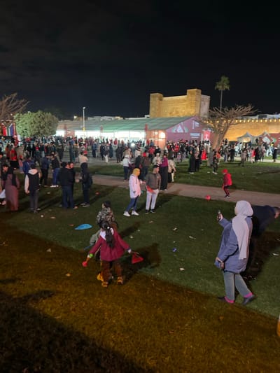 A night scene shows a crowd gathered near a building with a tower. People stand on a grassy area, some wearing hats and flags. A few individuals walk or stand in the foreground on the right, where debris litters the ground. The tower is illuminated, and trees are visible in the dark sky. The scene implies a public event or celebration.