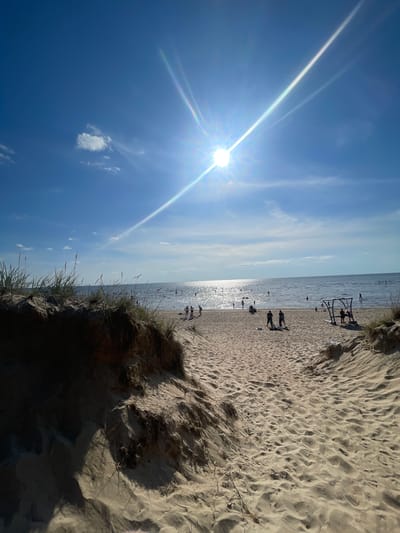 The image captures a view of a sandy beach leading towards the Gulf of Finland. The sun, a bright orb with visible lens flare, shines from the upper center of the frame, casting a strong glare on the water and illuminating the sandy path. The sky is a clear blue with a few wispy white clouds.

The foreground is dominated by sand dunes covered in dry grass, with a path of rippled sand winding towards the sea. Several people are visible on the beach and in the shallow water, some appearing to be walking, standing, or engaged in activities. In the distance, the water is calm and reflects the sunlight. A small, metallic playground structure with a roof is situated on the sand to the right.

The scene appears to be a public beach in Sosnovy Bor, Russia, on a sunny day, likely during the summer season, given the presence of people enjoying the water and beach.