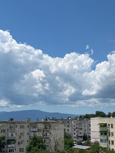 The image depicts a view of residential buildings in the city of Sopot, Bulgaria, under a bright blue sky with large, fluffy white clouds. In the foreground, multiple apartment buildings, some appearing aged with visible signs of wear, are arranged side-by-side. These buildings feature multiple stories with numerous windows, balconies, and protruding chimneys. Lush green trees are interspersed between and in front of the buildings. In the background, a range of blue-hued mountains forms the horizon. The overall scene suggests a residential urban area with natural mountainous scenery in the distance, under a partly cloudy sky.