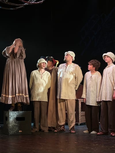 The image displays a theatrical performance on a stage, likely within a cultural center or theatre, located in Novoshakhtinsk, Russia. Six individuals, dressed in period-style costumes, are arranged across the stage. On the far left, a female figure in a long, flowing brown dress with ruffles stands atop a dark, cuboid stage prop, her hands covering her face in an gesture of distress. To her right, a male youth wearing a light beige tunic with laced neckline, dark trousers, and a light cap looks upwards and to the right. Adjacent to him, a partially obscured individual in a dark robe and a multicolored jester's hat is visible. At the center-right, a male adult, clad in a light beige tunic with laced neckline, matching trousers, and a light cap, stands with dark gladiatorial sandals, gazing upwards and to the right. To his immediate right, a male youth with dark, curly hair, dressed in a light beige tunic and dark trousers with light slippers, looks left towards the central adult. On the far right, a final male youth, wearing a light beige tunic, dark trousers, and a light cap, also looks upwards and to the right, wearing dark sandals. The stage features a dark background with abstract, branch-like suspended elements overhead and faint geometric patterns on the back wall, along with a partial wooden furniture prop on the right.