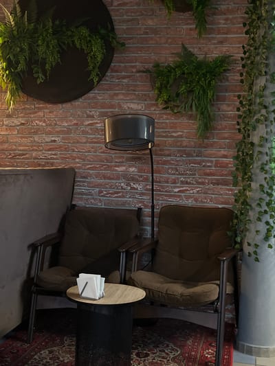 The image shows a lounge area with two brown armchairs, a small round side table with napkins, and a dark mesh-like container next to the table. A floor lamp with a grey shade stands between the chairs. The backdrop is a brick wall adorned with hanging plants and greenery. A patterned rug is partially visible in the corner. The location is likely a cafe or waiting area, potentially in Neftekamsk, Russia, though no specific details in the image confirm this. There are no people visible.