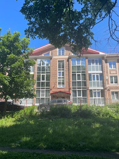 This is a daytime, eye-level, vertical, medium-shot photograph of a modern brick apartment building. The building has a red-tiled roof with white trim. The facade is primarily made of red brick with large, white-framed windows, some of which are floor-to-ceiling. The windows reflect the surrounding trees and sky. The building appears to be several stories high. In front of the building, there is a black metal fence and lush green grass and bushes. Two cars are partially visible behind the fence, one dark gray and one reddish-brown. The sky is clear and blue. The scene is likely a residential area, possibly a courtyard or street in a city. Based on architectural style, it is likely a newer construction in Vologda, Russia.