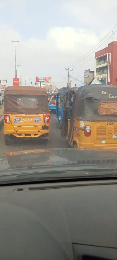 The image captures a static view of a multi-lane urban road in Akure, Nigeria, observed from the interior of a vehicle, as evidenced by the dashboard and windshield in the foreground. Multiple motorized tricycles, commonly referred to as Keke NAPEPs or auto-rickshaws, are stationary in traffic. The vehicle license plates, such as "AKR XXXX" visible on a tricycle, confirm the Akure location.
In the left lane, a yellow Piaggio-branded tricycle with a brown canopy is positioned directly ahead of the camera vehicle. Its rear displays "Age City NMT" and illuminated red brake lights. In the adjacent right lane, a yellow TVS-branded tricycle with a black canopy, marked "De Easy" on its rear, is also stationary; its right side panel is open. Occupants are visible within the tricycles but are not individually identifiable.
Further down the road, additional tricycles and a light blue sedan are present, indicating a queue of vehicles. Overhead streetlights and utility poles line the road, with power lines crisscrossing the upper frame. Traffic lights are visible in the distance, one displaying a green signal. On the right side of the road, a multi-story building with distinct red and white facades and balconies stands. Various commercial billboards are discernible in the background, including one advertising "Love or Veggies" and another displaying "734M". The sky overhead is predominantly overcast with patches of brighter cloud.