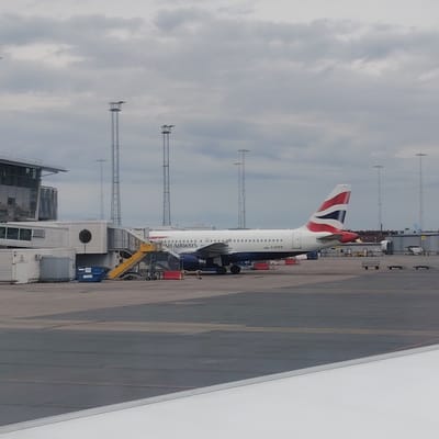 The image displays an airport apron scene at Stockholm-Arlanda Airport in Sweden, captured under an overcast sky. A British Airways Airbus A320-200 aircraft, identified by the registration G-EUPN on its fuselage, is centrally positioned and stationary on the tarmac. The aircraft is predominantly white with a blue underbelly and features the "BRITISH AIRWAYS" livery, including the distinct red, white, and blue "Speedmarque" on its tail fin.
A yellow mobile passenger stairs unit is positioned at the forward left door of the aircraft, with red safety barriers or cones marking the immediate area. To the left of the aircraft, a segment of an airport terminal building is visible, characterized by horizontally ridged paneling and a prominent circular window or opening on one of its connecting structures. A large blue dumpster is situated near this terminal section and the aircraft's stairs.
The background features several tall, metallic lattice-style light poles equipped with platforms, typical of airport lighting infrastructure. Further back, to the right of the aircraft's tail, various airport buildings are present, including one with a large, cylindrical structure resembling an engine test cell or maintenance facility. The tail section of another distant aircraft is partially visible behind the light poles. The foreground includes a white, unidentifiable smooth surface, likely a wing or a window frame, partially obscuring the lower right portion of the image. The ground is a dark grey paved tarmac with visible markings and seams. No discernible individuals are clearly visible.