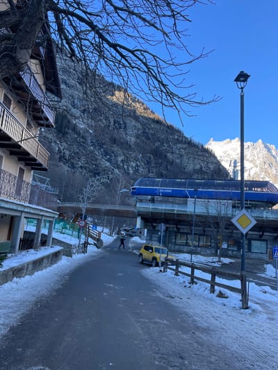 A vibrant winter scene in Courmayeur, Italy, depicts a clear, sunny day in a mountain town. The image, oriented as if viewed looking up a street towards a mountain, features a steep, rocky mountain face dominating the background, with sunlit, snow-capped peaks visible further up.

In the mid-ground, a prominent blue gondola station, labeled "DOLONNE," runs parallel to the street, suggesting a ski resort or mountain transport hub. Below it, a paved road, partially covered with snow and ice, stretches from the foreground into the distance. A yellow jeep-like vehicle is parked on the right side of the road, and a single person walks along the icy pavement in the mid-ground. A wooden fence lines the roadside.

To the left, a bare-branched tree occupies the immediate foreground, its intricate branches silhouetted against a brilliant clear blue sky. Partially visible buildings with balconies, typical of alpine architecture, are located on the left and right sides of the foreground and mid-ground.

Notable objects include a street lamp, a yellow diamond-shaped road sign indicating a priority road, a blue square sign for a pedestrian crossing, and a circular sign with a "2.1m" height limit. The clear sky and strong shadows suggest it is daytime, likely morning or early afternoon, with cold, crisp weather conditions conducive to snow and ice. The overall setting is that of an active, functional mountain resort town during the winter season.
